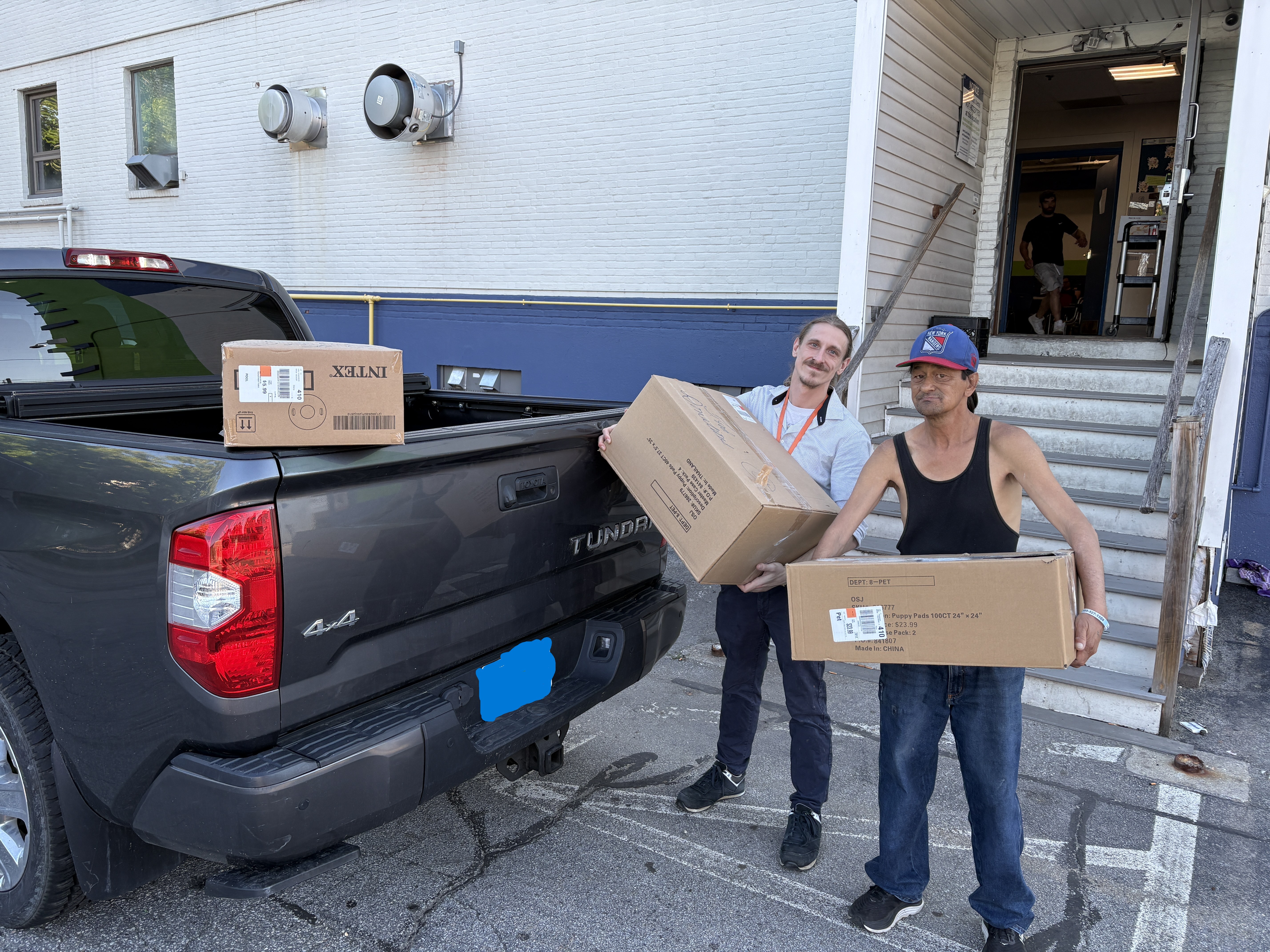 Two people carrying large boxes beside a pickup truck outside a community building
